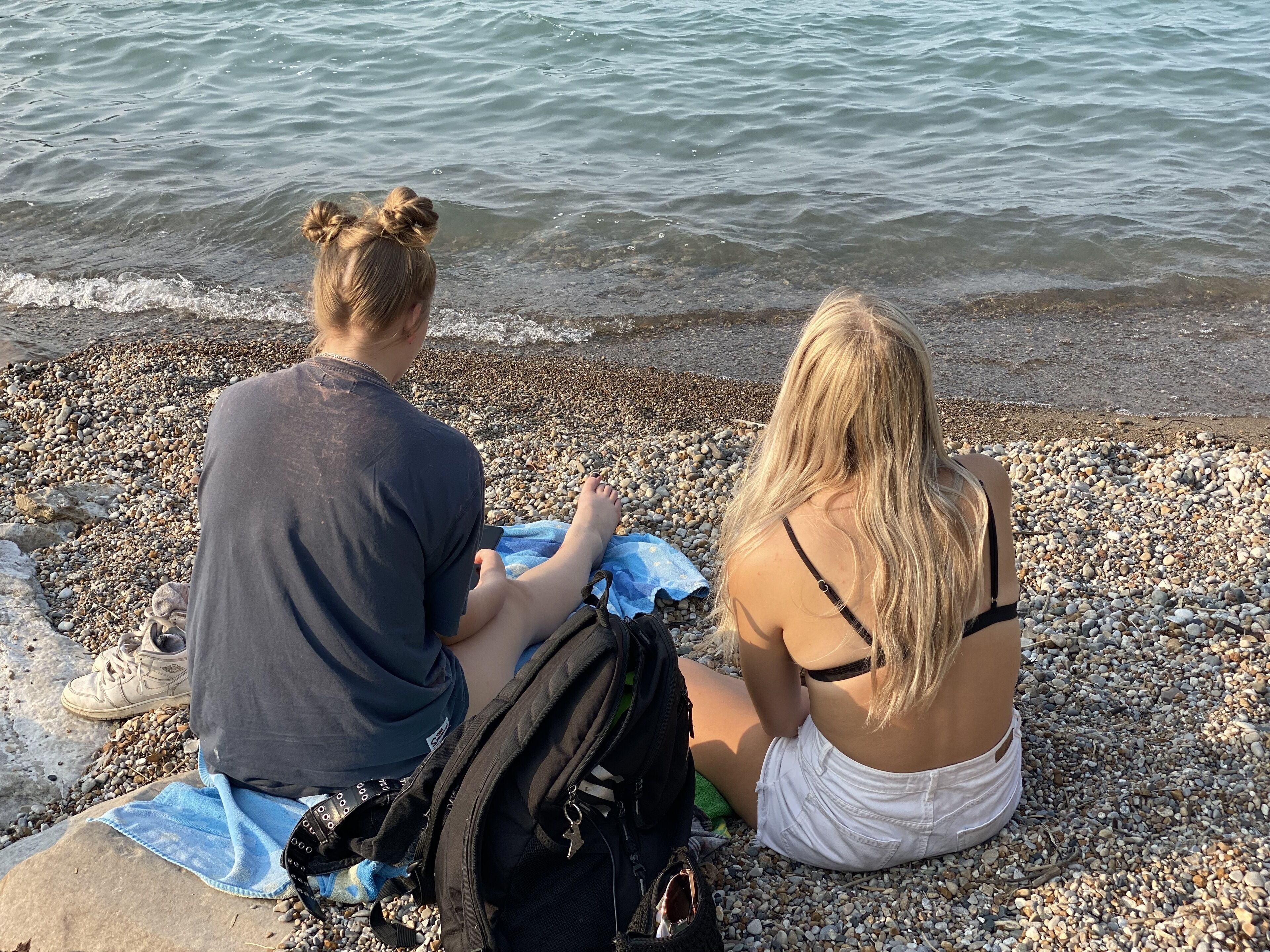 Two people sitting on a pebbled beach with a body of water in the background.
