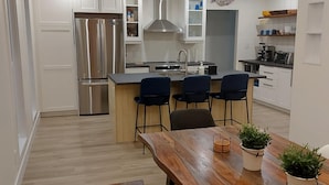Una cucina moderna con mobili bianchi, un frigorifero in acciaio inossidabile, un'isola centrale con sgabelli da bar e un tavolo da pranzo in legno con piante in vaso.