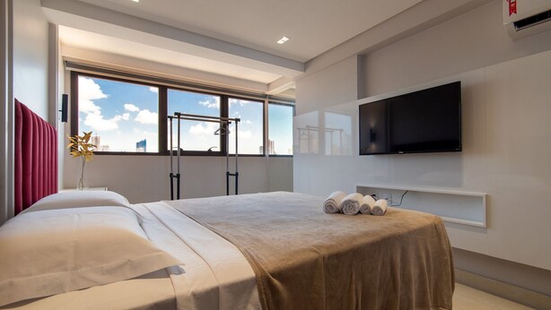 A modern hotel room with a large bed, a TV mounted on the wall, and a window with a city view. - Incrível Suíte no Manaíra Apart Hotel (João Pessoa)