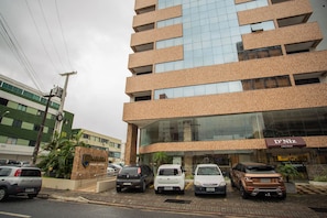 A multi-story hotel with a modern design, featuring a prominent glass facade and a parking area with several cars. - Incrível Suíte no Manaíra Apart Hotel (João Pessoa)