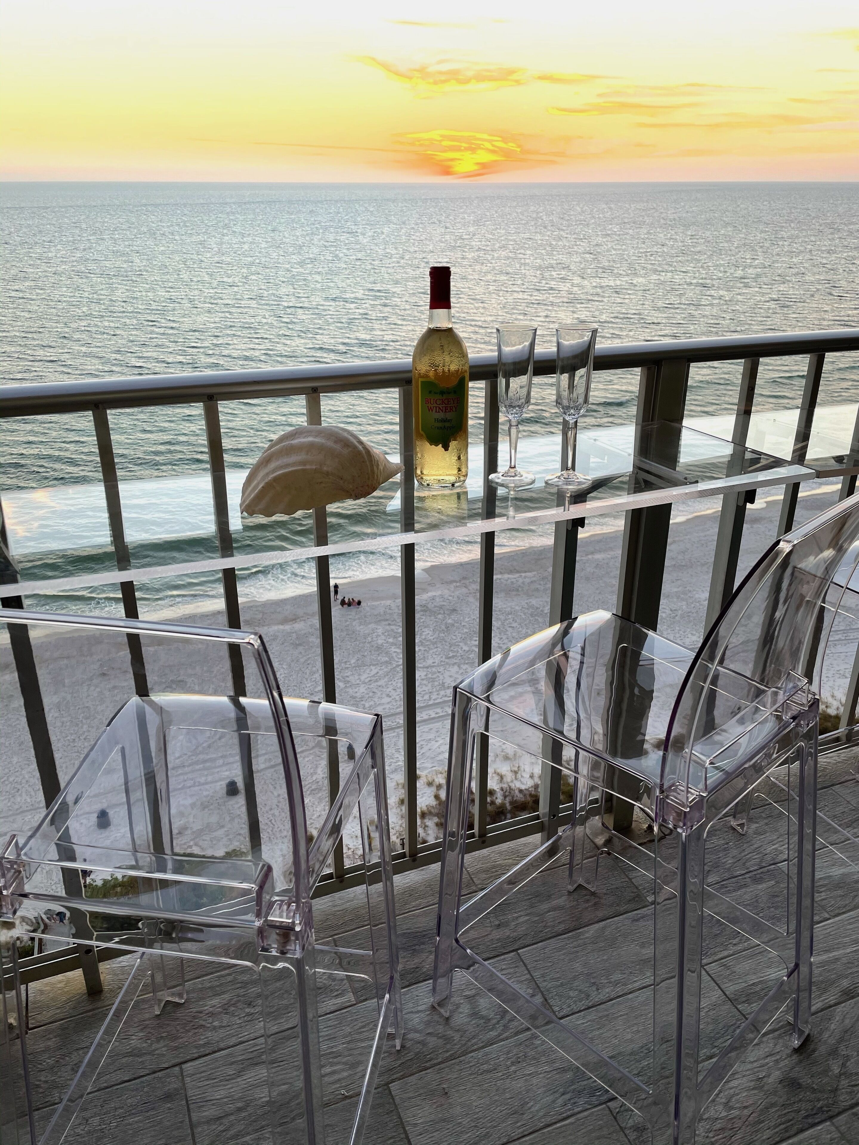 En balkon med glasstole, en flaske Buckeye Winery vin og et glas på gelænderet med udsigt til havet i solnedgang.