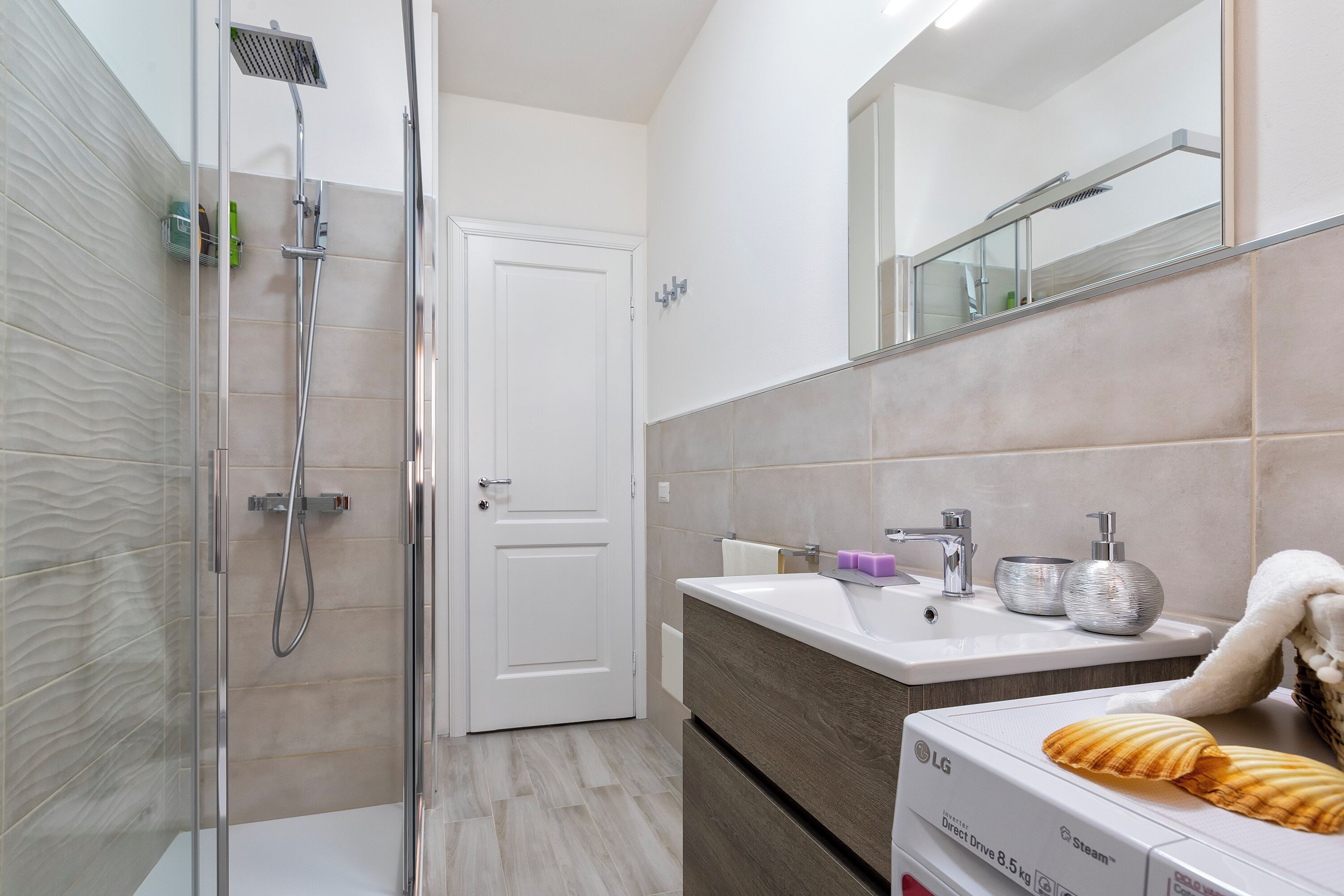 A modern bathroom with a glass shower enclosure, a white sink with a mirror above, and a beige tiled wall.