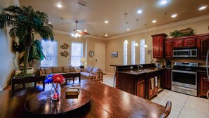 A spacious living room with a large wooden dining table, a comfortable sofa, and a kitchen area with wooden cabinets and stainless steel appliances. - Lovely 2-bedroom unit with a pool & workspace (South Padre Island)