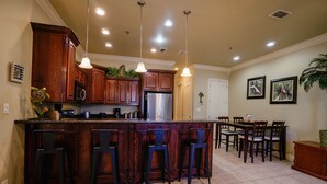 A kitchen with wooden cabinets, a stainless steel refrigerator, a bar area with stools, pendant lighting, and a dining area with chairs and a table. - Lovely 2-bedroom unit with a pool & workspace (South Padre Island)