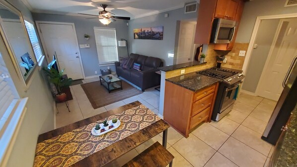 A compact kitchen with a stove, microwave, and cabinets. A dining table with a patterned table runner and potted plants is in the foreground. A living room with a sofa and a cityscape painting is visible in the background.