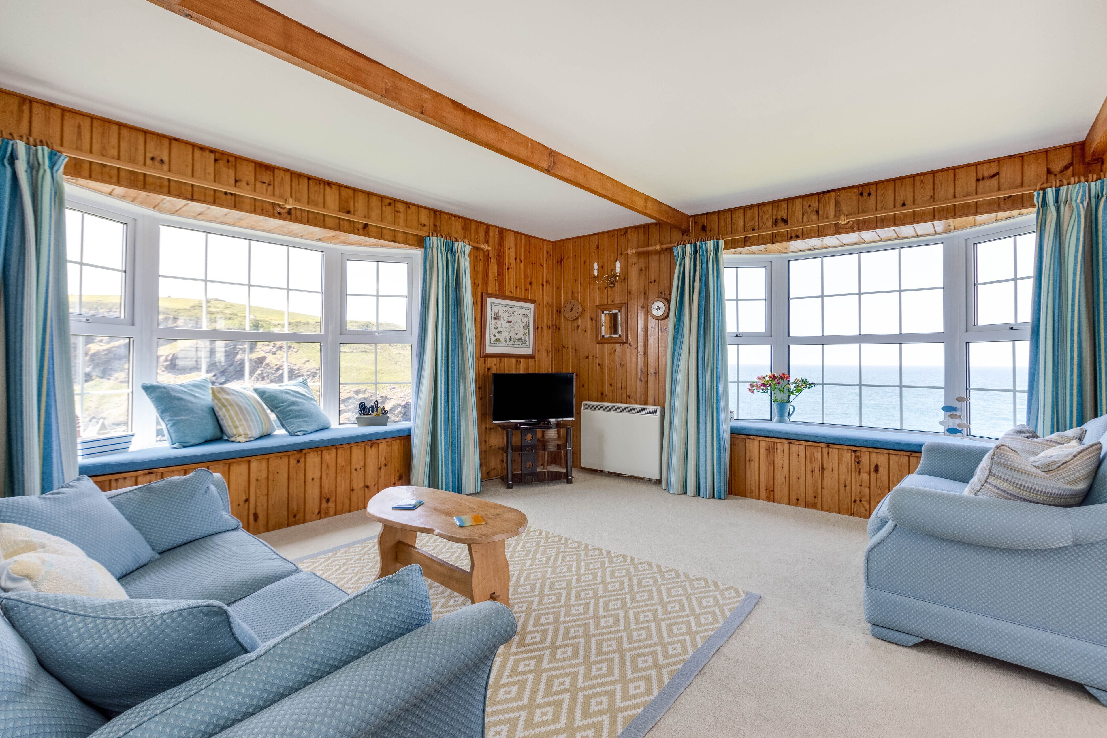 Una habitación acogedora con paredes de madera, ventanas amplias con cortinas, un sofá, una mesita y un televisor.