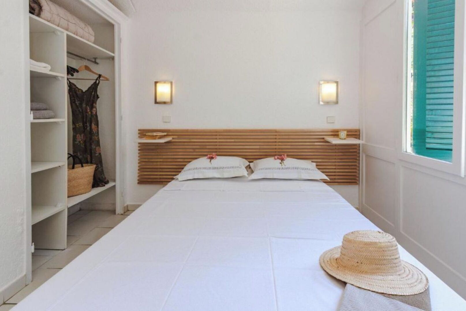 A bedroom with a wooden headboard, two bedside lamps, a bed with white linens, a wardrobe with shelves, and a window with green shutters.