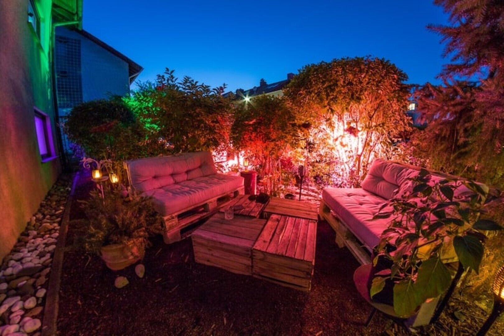 Un espace extérieur aménagé avec deux canapés et une table basse, entouré de plantes et d’arbres, le tout éclairé par des jeux de lumière colorés.