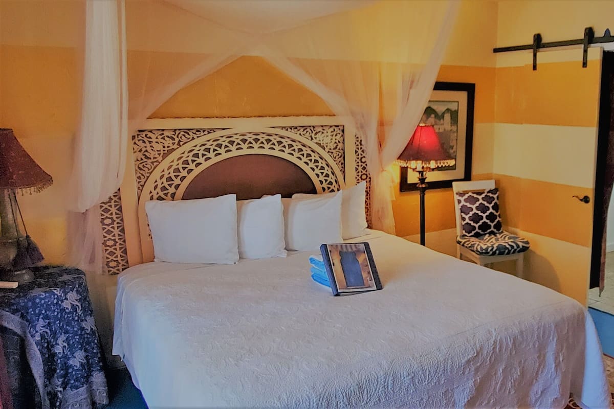 Ein Bett mit Baldachin, ein Nachttisch mit Lampe, ein Sessel mit gemustertem Kissen und ein durchgehender Türspalt sichtbares Badezimmer.