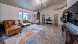 A spacious living room with a sectional sofa, a coffee table, and a large rug. There is a television on a wooden stand and a bookshelf with decorative items.