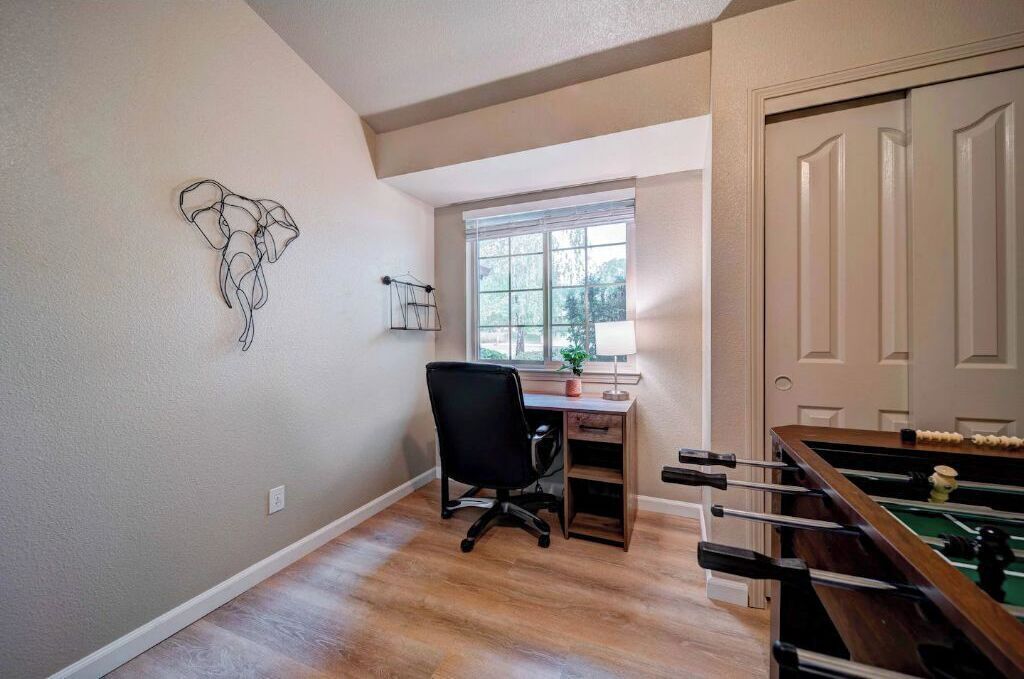 Une pièce avec un bureau en bois, un fauteuil de bureau noir, une table de baby-foot et une fenêtre donnant sur des arbres.