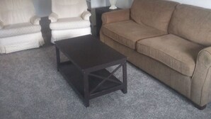 Un salon avec un canapé beige, deux fauteuils blancs, une table basse en bois foncé, une table d’appoint blanche avec une lampe, et un tableau encadré accroché au mur.