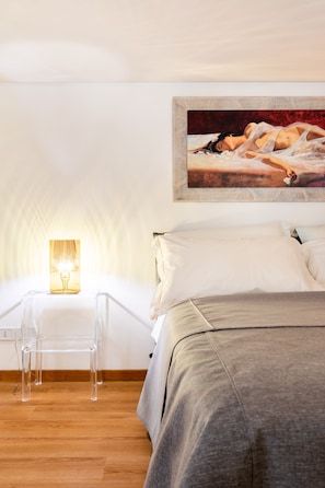 A bedroom with a bed, a bedside table with a lamp, a clear chair, and a painting on the wall. - Rotonda 23 - Loft in Pantheon (Roma)