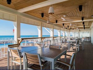 Ein überdachter Außenbereich zum Essen mit Tischen und Stühlen, großen Fenstern und Blick aufs Meer.