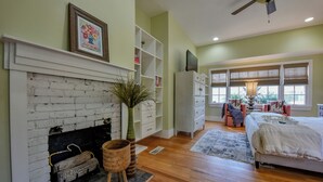 A bedroom with a fireplace, wooden flooring, a bed, a TV, and a window with blinds.