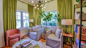 A cozy living room with a pink armchair, a white sofa, and a green ottoman. There are two windows with green curtains, a large plant, and a bookshelf with books.