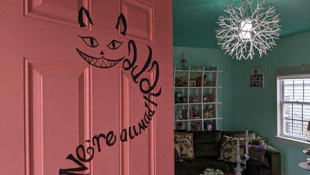 A room with a pink door featuring a cat face and the text "We're always here." A white chandelier hangs from the ceiling, and a bookshelf filled with various items is visible. - Escape to Wonderland (Oklahoma City)