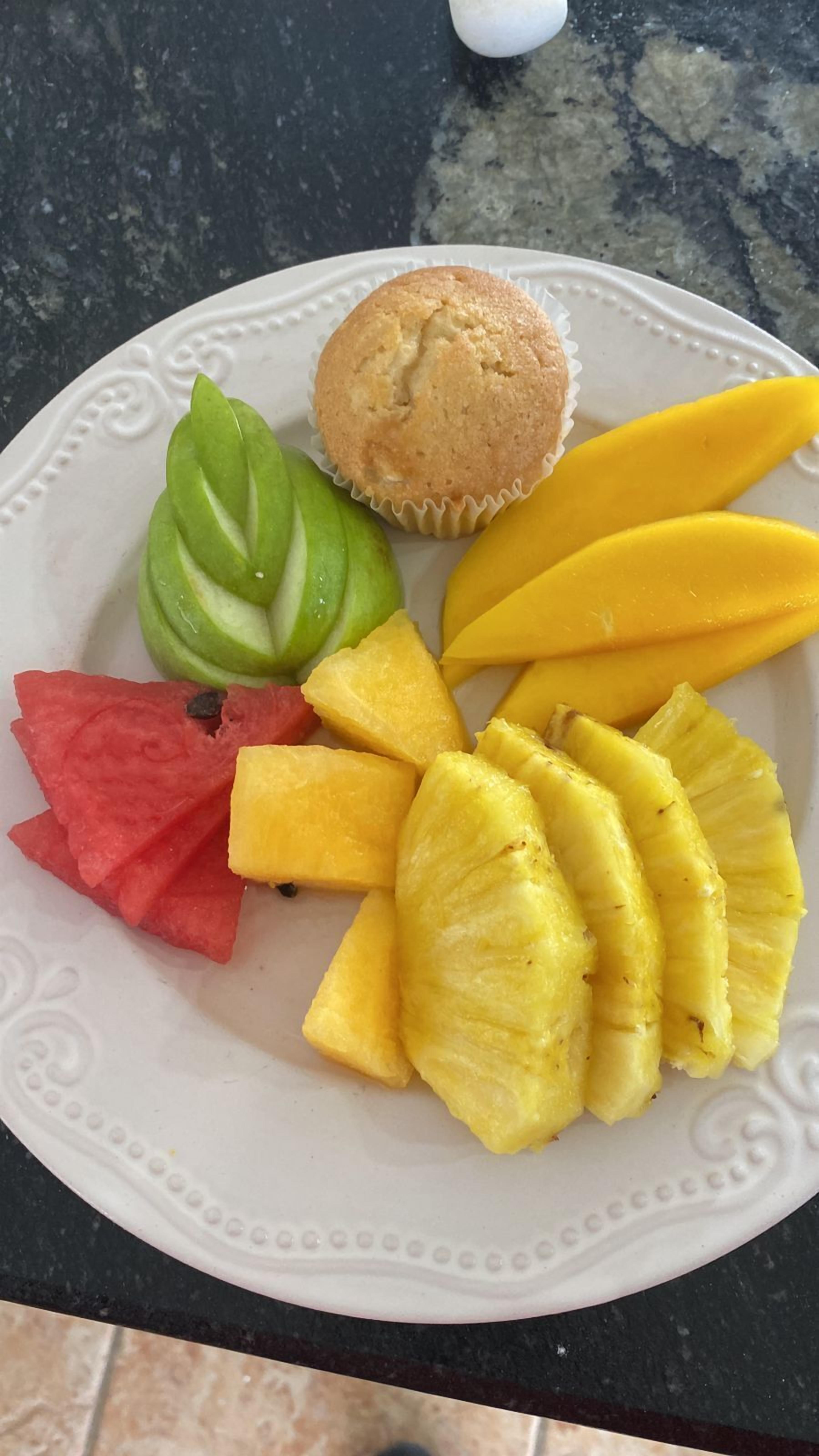 Un piatto con un muffin, fette di mango, ananas, anguria e mele a spicchi.