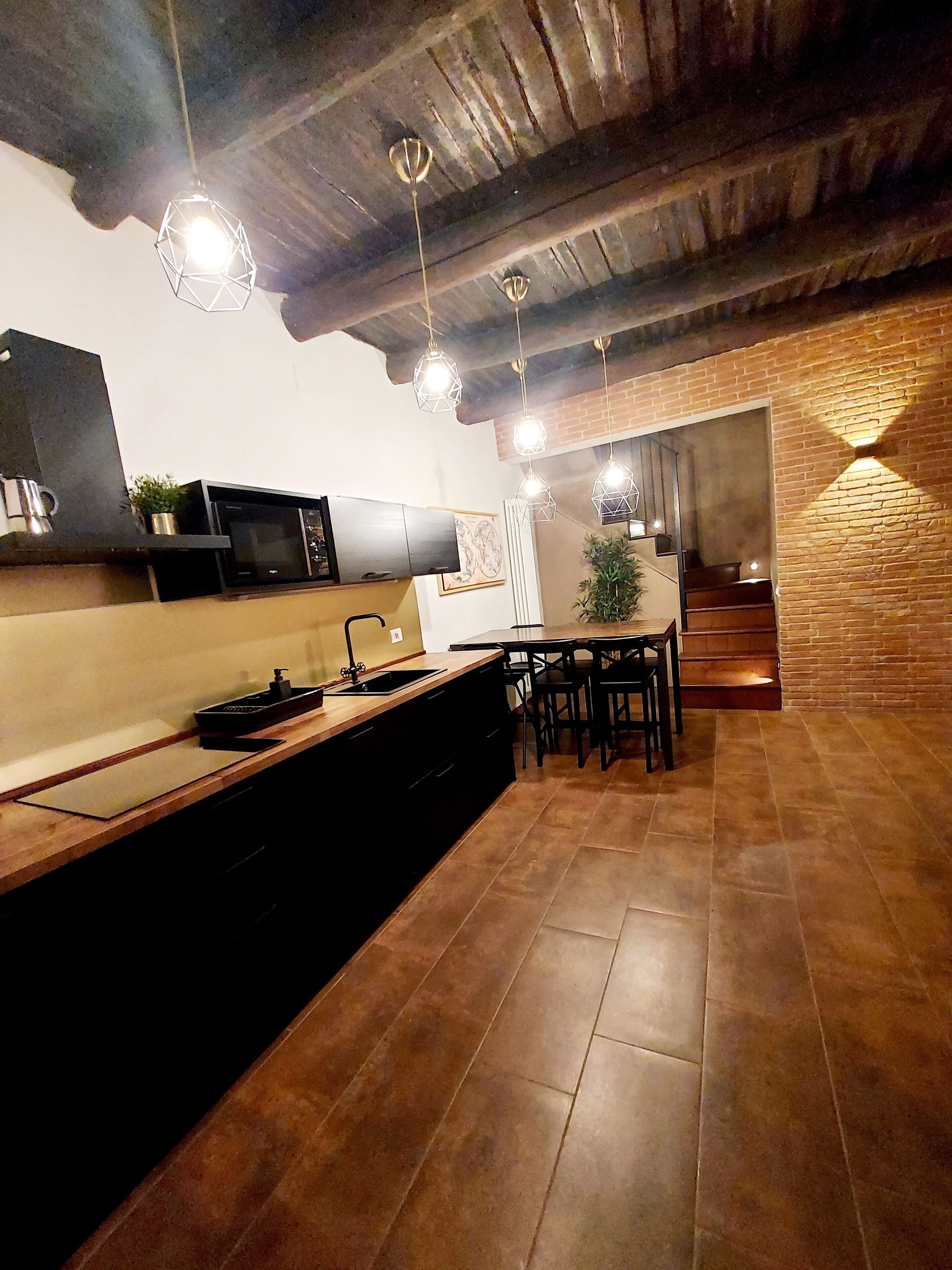 A modern kitchen with a central island, black cabinetry, and a microwave. There are hanging pendant lights and a staircase leading to an upper level.