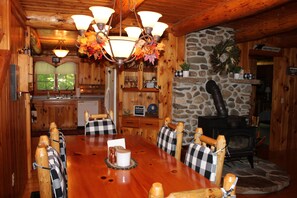 A wooden dining room with a large table, checkered chairs, a chandelier, a stone fireplace, and a kitchen area with wooden cabinets and a sink. - Dog-Friendly 5 Bedroom Log Cabin with Hot Tub in the Heart of Southern Maine  (Brownfield)