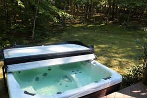 A hot tub with a black lid, surrounded by a wooden deck, set in a grassy area with trees in the background. - Dog-Friendly 5 Bedroom Log Cabin with Hot Tub in the Heart of Southern Maine  (Brownfield)