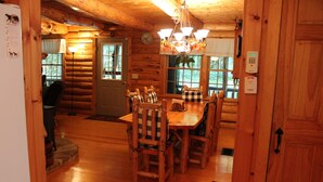 Interior de uma cabana de madeira com sala de jantar, cadeiras xadrez e mesa de madeira.