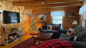 Interior acolhedor de uma cabana, com decoração em madeira, uma árvore de Natal, um sofá e uma televisão.