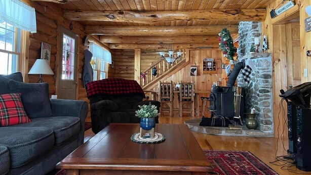 Interior acolhedor de uma cabana de toras, com lareira, escada e sala de estar com sofá e mesa de centro.