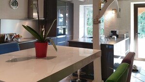 A modern kitchen with a white countertop, a red potted plant, and a dining area with colorful chairs. - Au Fil des Hirondelles (Bio)