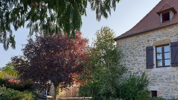 A stone building with a red roof, surrounded by a gravel driveway and various trees. - Au Fil des Hirondelles (Bio)