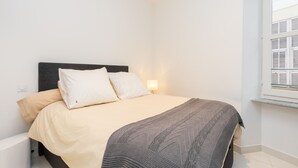 A bedroom with a bed, white walls, a window with shutters, and a bedside lamp.