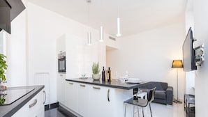 A modern kitchen with white cabinets, a black countertop, and stainless steel appliances. There is a bar stool and a grey sofa in the living area.