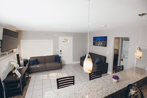 A modern living room with a grey sofa, a dining area with a granite countertop, and a kitchen area with a built-in oven.