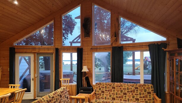 A cozy cabin interior with a wooden ceiling, large windows, a sofa, a table, and a chair.