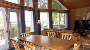 A wooden dining area with a large table and chairs, a television, and large windows overlooking a deck.
