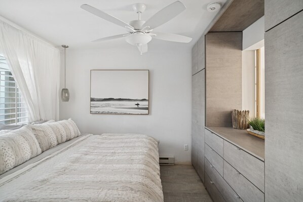 Un dormitorio con cama, ventilador de techo, ventana con cortinas, una obra de arte en la pared y un banco empotrado con planta.