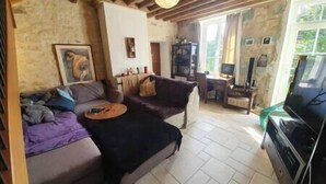 A cozy living room with a sofa, a coffee table, and a TV. There is a fireplace and a window with curtains. - Cozy Cottage (Éméville)