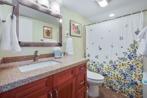 A bathroom with a granite countertop sink, wooden cabinets, a white toilet, and a shower curtain with a floral pattern.