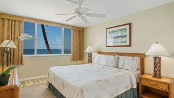 A hotel room with a large bed, two bedside lamps, a ceiling fan, a window with a view of the ocean, and a framed picture on the wall.