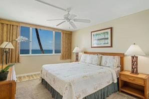 A hotel room with a large bed, two bedside lamps, a ceiling fan, a window with a view of the ocean, and a framed picture on the wall.