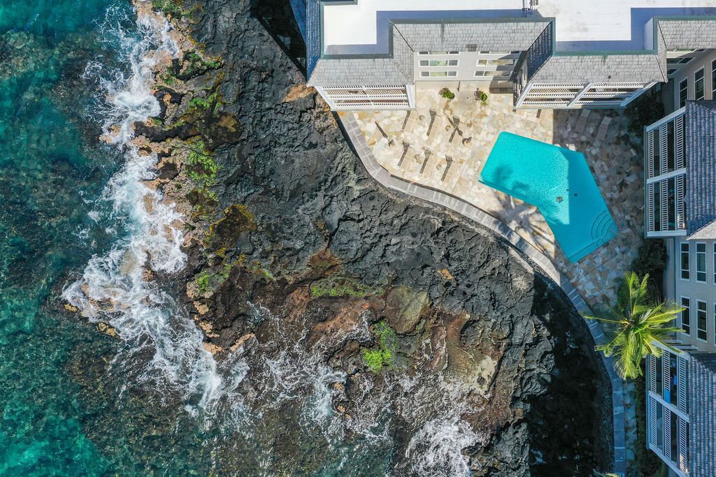 An aerial view of a coastal property with a swimming pool, rocky shoreline, and a building with a white facade and grey roof.
