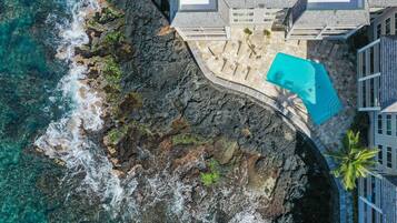 An aerial view of a coastal property with a swimming pool, rocky shoreline, and a building with a white facade and grey roof.