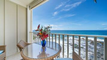 A balcony with wicker furniture, a glass table, and a vase of flowers. The view includes the ocean and rocky shoreline.