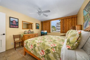 A bedroom with a bed, a desk, a chair, a ceiling fan, a window with a view of palm trees, and a painting on the wall.