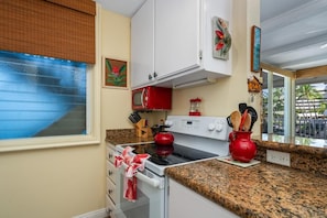 Eine Küche mit Herd, Backofen und Mikrowelle. Ein Fenster mit Jalousien, ein Bild an der Wand und Blick auf Palmen draußen.