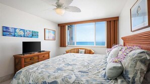Un dormitorio con una cama, un televisor sobre una cómoda de madera, un ventilador de techo, una ventana con cortinas y obras de arte en la pared.