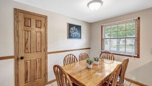 A dining area with a wooden table, six chairs, a potted plant, a framed sign, a wooden door, and a window with blinds. - Shadow Lane Tailgate House - Newly Remodeled (Green Bay)
