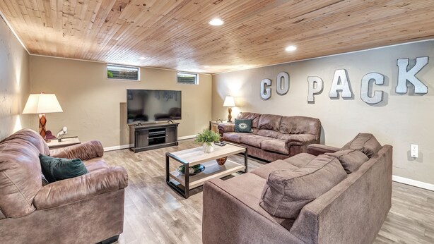 A cozy living room with a wooden ceiling, two leather sofas, a coffee table, a TV, and a 'GO PACK' sign on the wall. - Shadow Lane Tailgate House - Newly Remodeled (Green Bay)