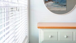 A room with a window with blinds, a dresser with drawers, and a round mirror on the wall. - Newport's favorite Neighborhood 3bed (Newport)