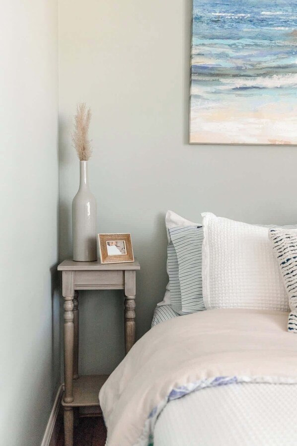 A bedroom with a bed, a nightstand with a vase and a picture frame, and a painting on the wall.